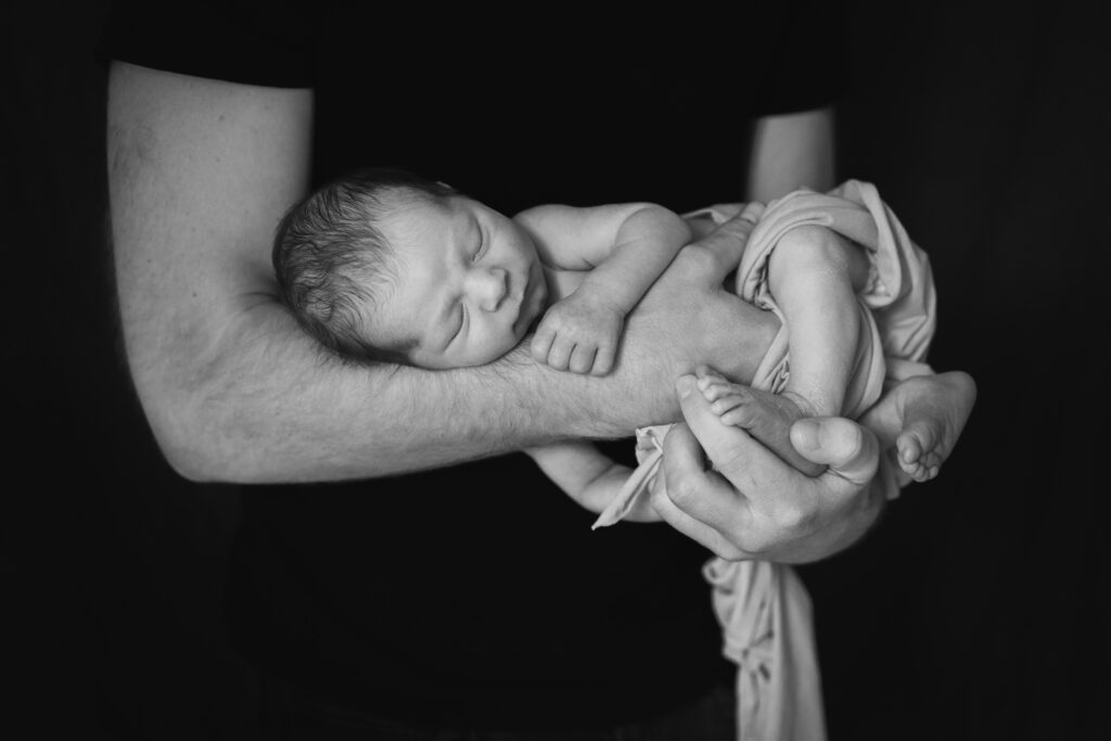Schwarz-weiß Bild eines neugeborenen Babies, das von den Armen des Papas gehalten wird.