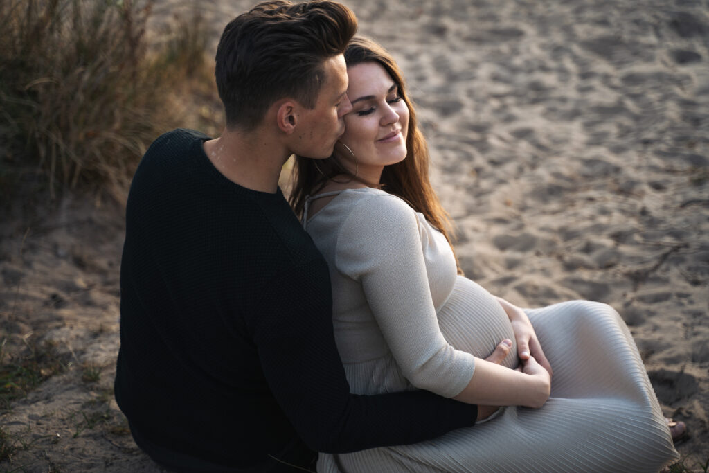 Eine schwangere Frau sitzt mit geschlossenen Augen an ihren Freund gelehnt im Sand.