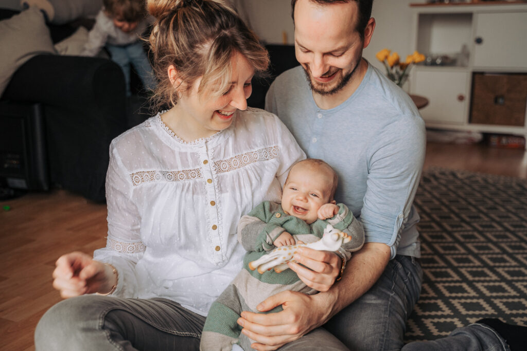 Lachendes Baby auf dem Arm seiner Eltern, während der Bruder im Hintergrund turnt.
